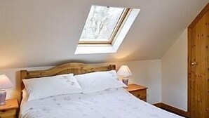Une chambre avec un puits de lumière, une tête de lit en bois, un lit avec une literie blanche, deux tables de chevet avec des lampes et une armoire en bois.