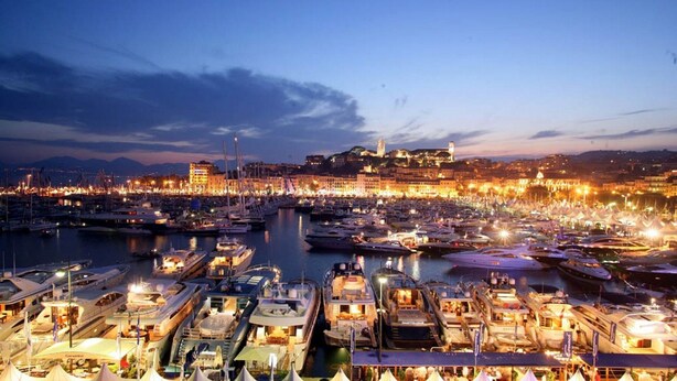 Un puerto deportivo al atardecer con numerosos barcos amarrados, un edificio destacado al fondo y un paisaje urbano a través del agua.