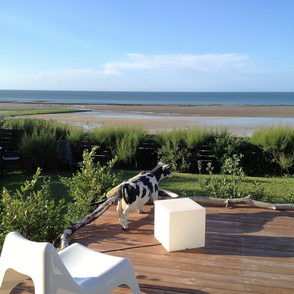 A deck with a white plastic chair and a white block, overlooking a beach with a cow statue in the foreground.