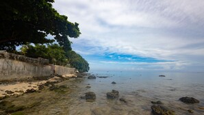 Ein Strand mit klarem, flachem Wasser, Felsen und einer Betonmauer mit Vegetation links.