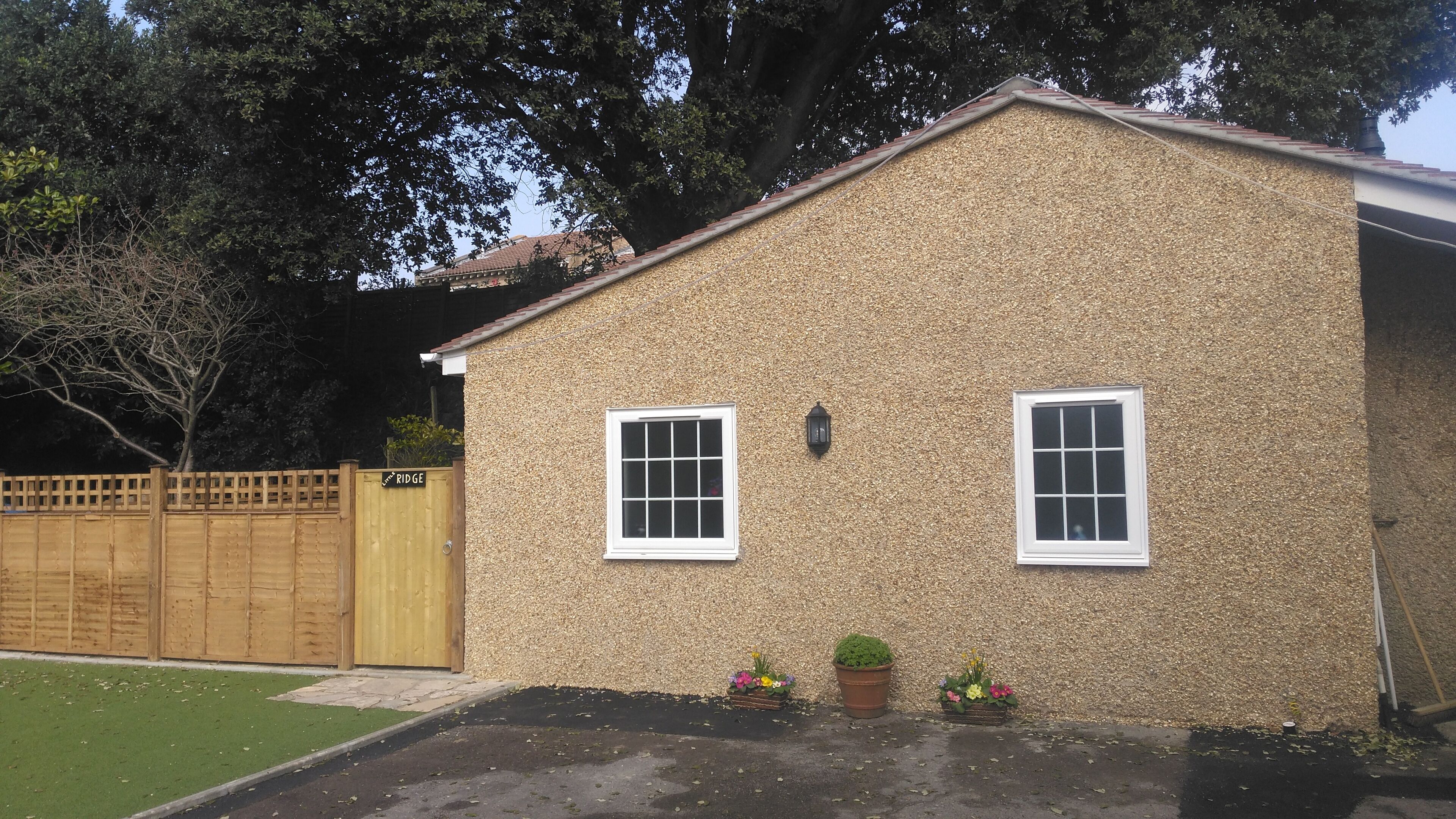 Une maison de plain-pied avec une façade à texture, deux fenêtres à cadre blanc, un petit porche agrémenté de plantes en pot et une clôture en bois.