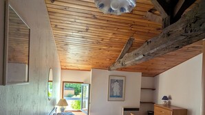 Ein Schlafzimmer mit Holzbalkendecke, einem Bett, einem Nachttisch mit Lampe, einer Kommode und einem Fenster mit Blick auf Grünflächen.
