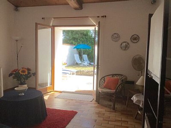 A room with a wooden floor, a red rug, a chair, a table with a vase of flowers, and a door leading outside.