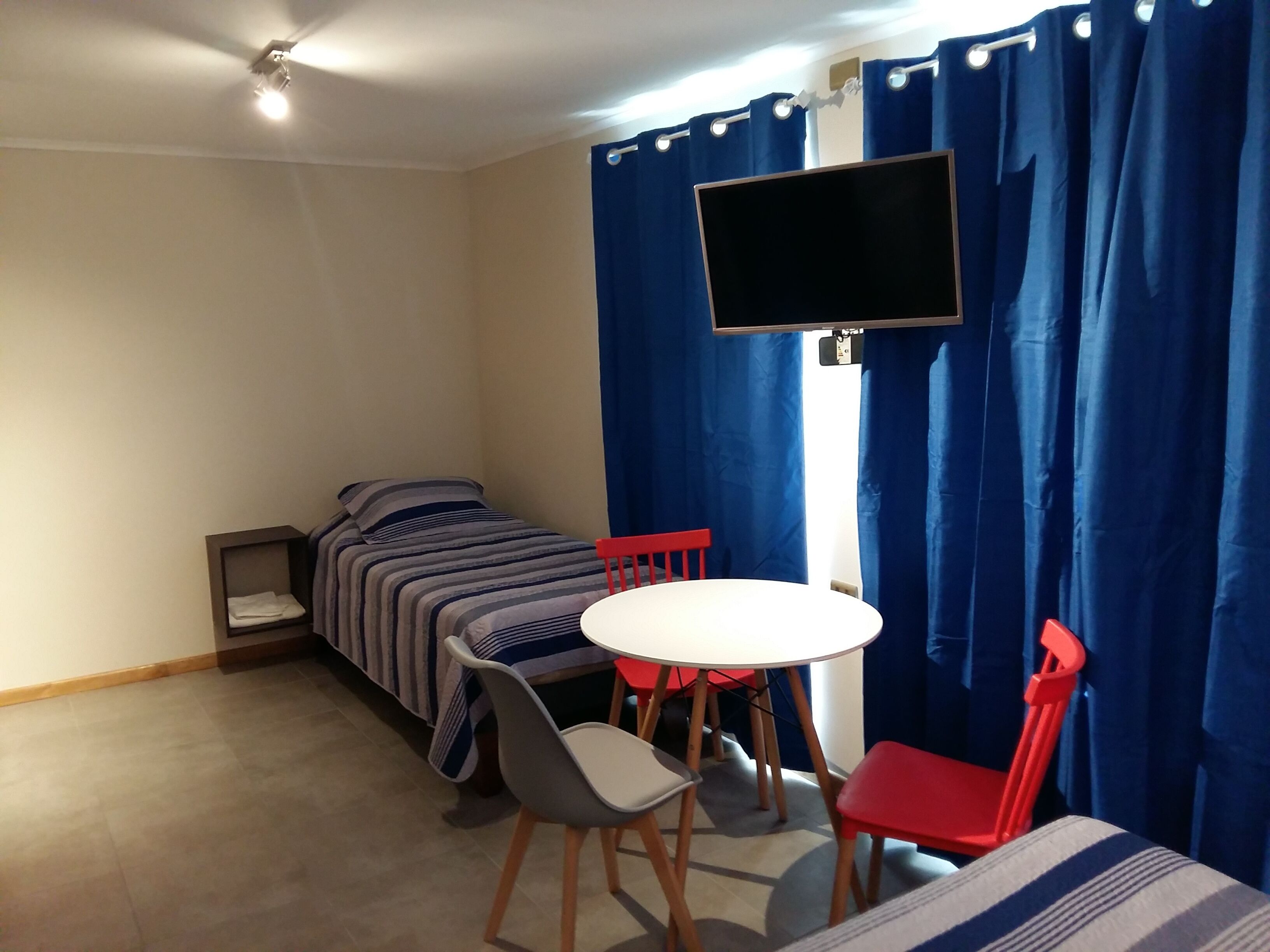 Una habitación pequeña con una cama individual, una mesita, dos sillas y un televisor fijado a la pared.
