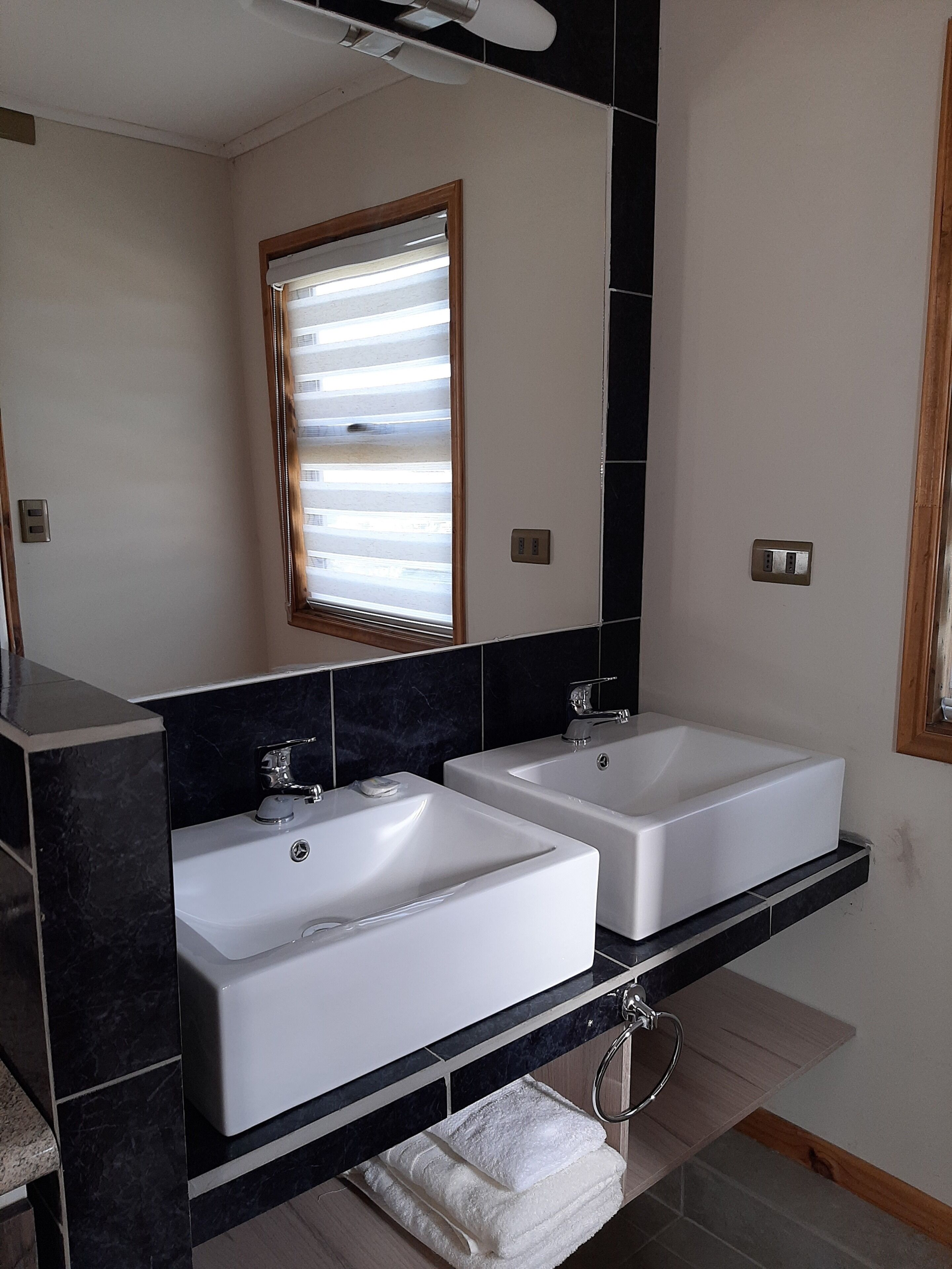A modern bathroom with two white sinks, a large mirror, and a window with blinds.