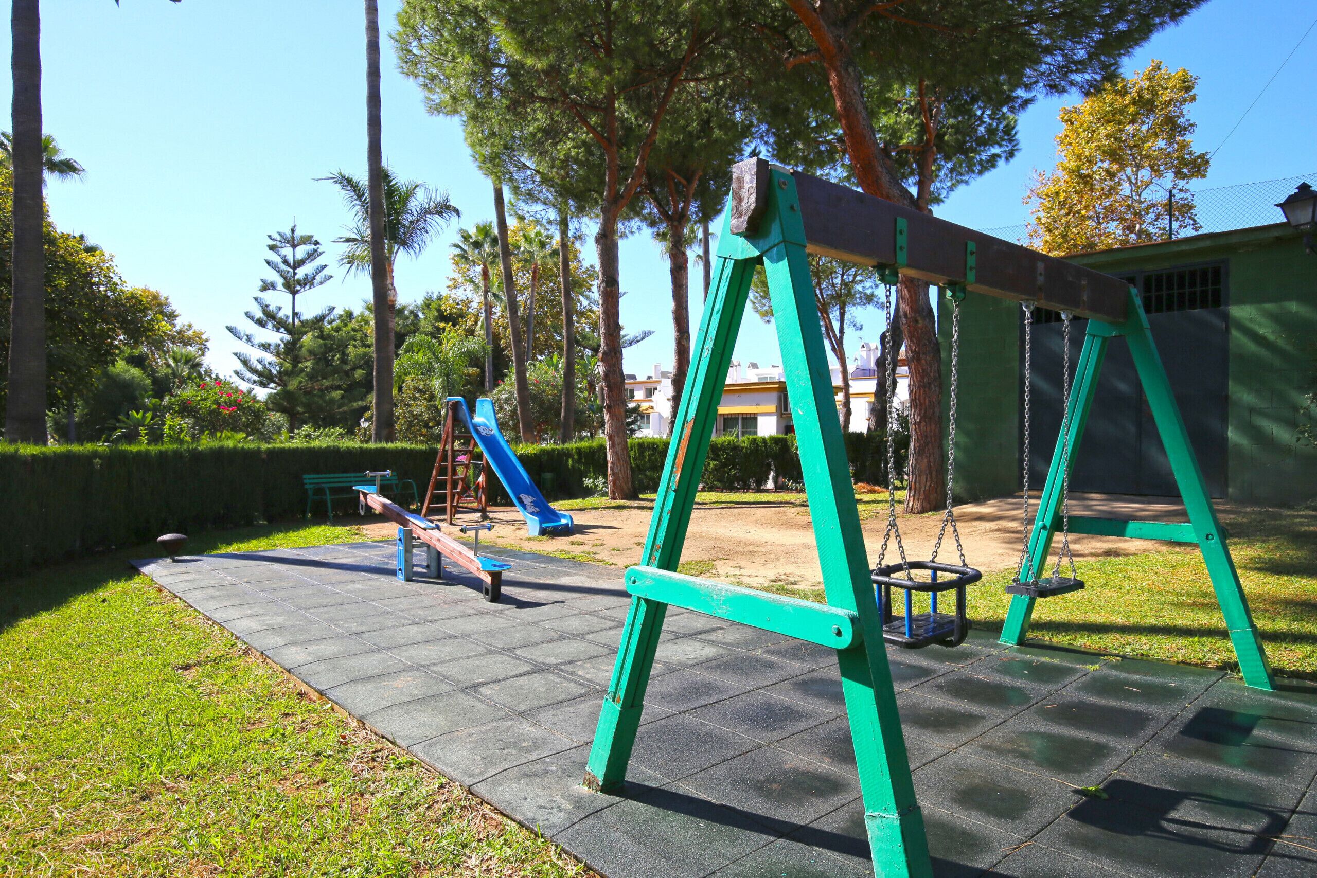 Un terrain de jeux avec des balançoires et un toboggan, entouré d’arbres et avec un bâtiment en arrière-plan.
