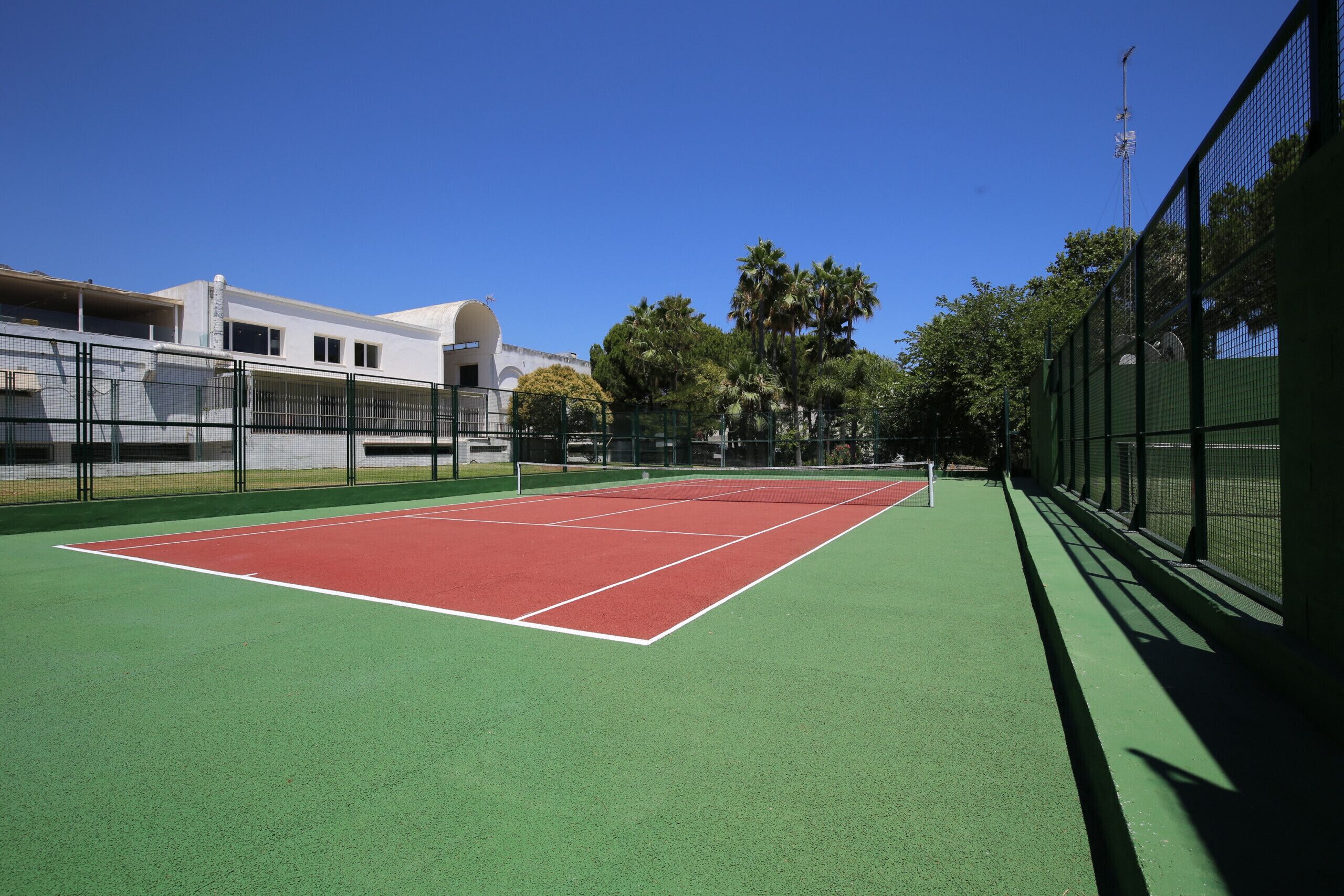 Una cancha de tenis con superficies verde y roja, rodeada por una valla, y un edificio con cúpula al fondo.