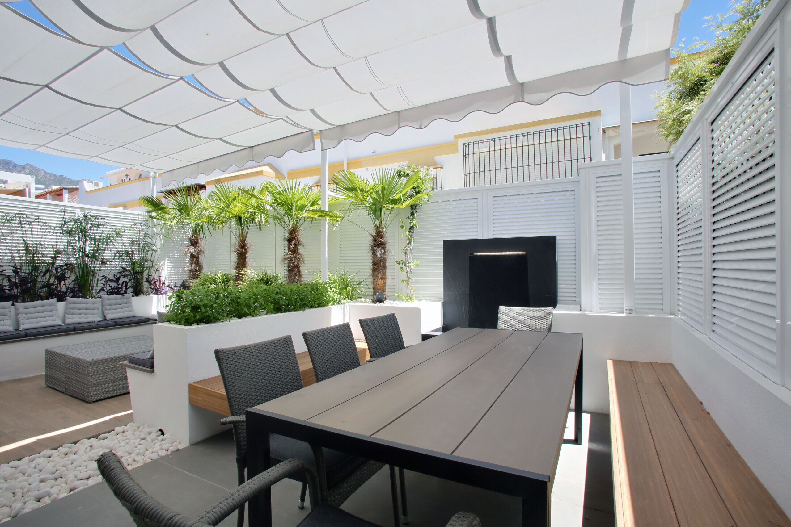 Un espacio comedor al aire libre con techo, con una mesa larga, sillas y un banco. Hay plantas y una zona de estar pequeña con cojines.