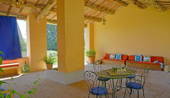 A covered patio with a round table, chairs, and a sofa with cushions.