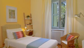 A bedroom with a bed, a chair, a bedside table, a lamp, a window with curtains, and a wall-mounted shelf with a plant.