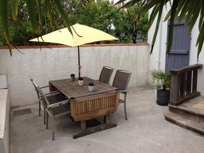Une table en bois pour l’extérieur, avec quatre chaises, un parasol jaune et des plantes en pot.
