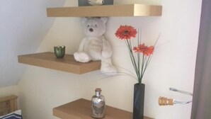 Un dormitorio con una cama, un cojín a rayas y tres estantes de madera. El estante superior sostiene un jarrón con flores, el estante central tiene un oso de peluche, y el estante inferior contiene una botella.