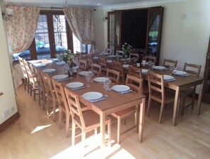 A dining area with multiple wooden tables and chairs, large windows with curtains, and a view of the outside.