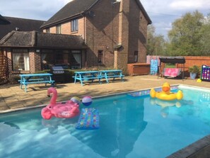 A backyard with a swimming pool, two blue picnic tables, a pink flamingo float, a yellow duck float, and a colorful inflatable pool toy.