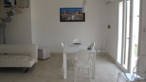 Una sala de estar moderna con un sofá blanco, una mesa de comedor con sillas, un cuadro en la pared y ventanas grandes.