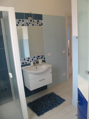 A bathroom with a white sink, blue and white tiles, a glass shower enclosure, and a dark blue rug.