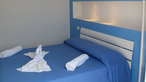 Una cama bien hecha con ropa de cama azul y toallas blancas, un cabecero blanco y una lámpara de pared.