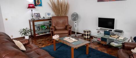 Ein Wohnzimmer mit braunen Ledersofas, einem Glastisch, einem Bücherregal, einem Ventilator und einem Fernseher.
