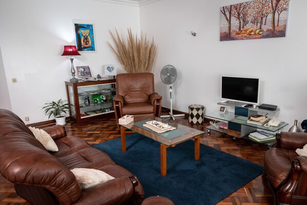 A living room with brown leather sofas, a glass coffee table, a bookshelf, a fan, and a TV. - TMO Residence 2 P. D.L (Ponta Delgada)