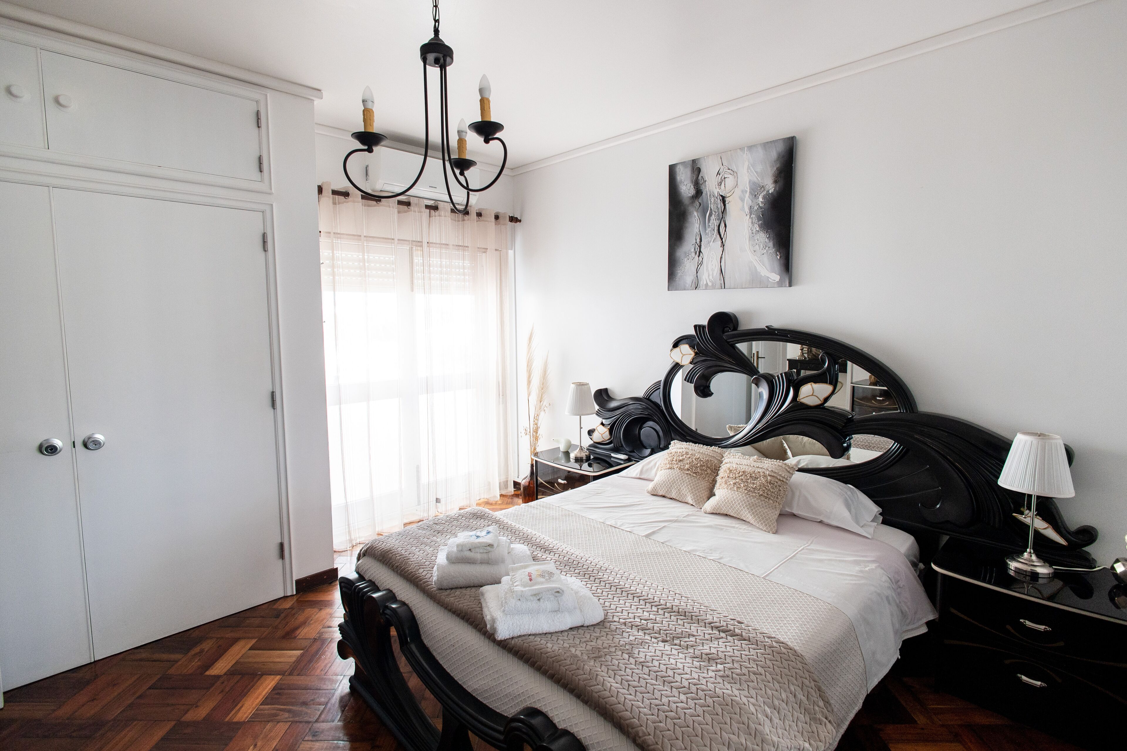 A bedroom with a large bed, a chandelier, a bedside table with a lamp, and a painting on the wall.