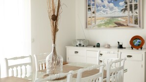 A dining area with a glass table, white chairs, a vase with branches, and a painting of a beach scene on the wall. - TMO Residence 2 P. D.L (Ponta Delgada)
