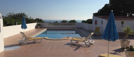 A rooftop pool area with two sun loungers and blue umbrellas, surrounded by a white wall and potted plants.