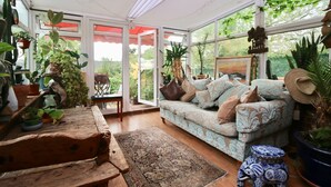 A conservatory with a patterned sofa, wooden furniture, and various potted plants. - Unique 18th Century village cottage near Loch Lomond and Trossachs/ shop and pub (Killearn)