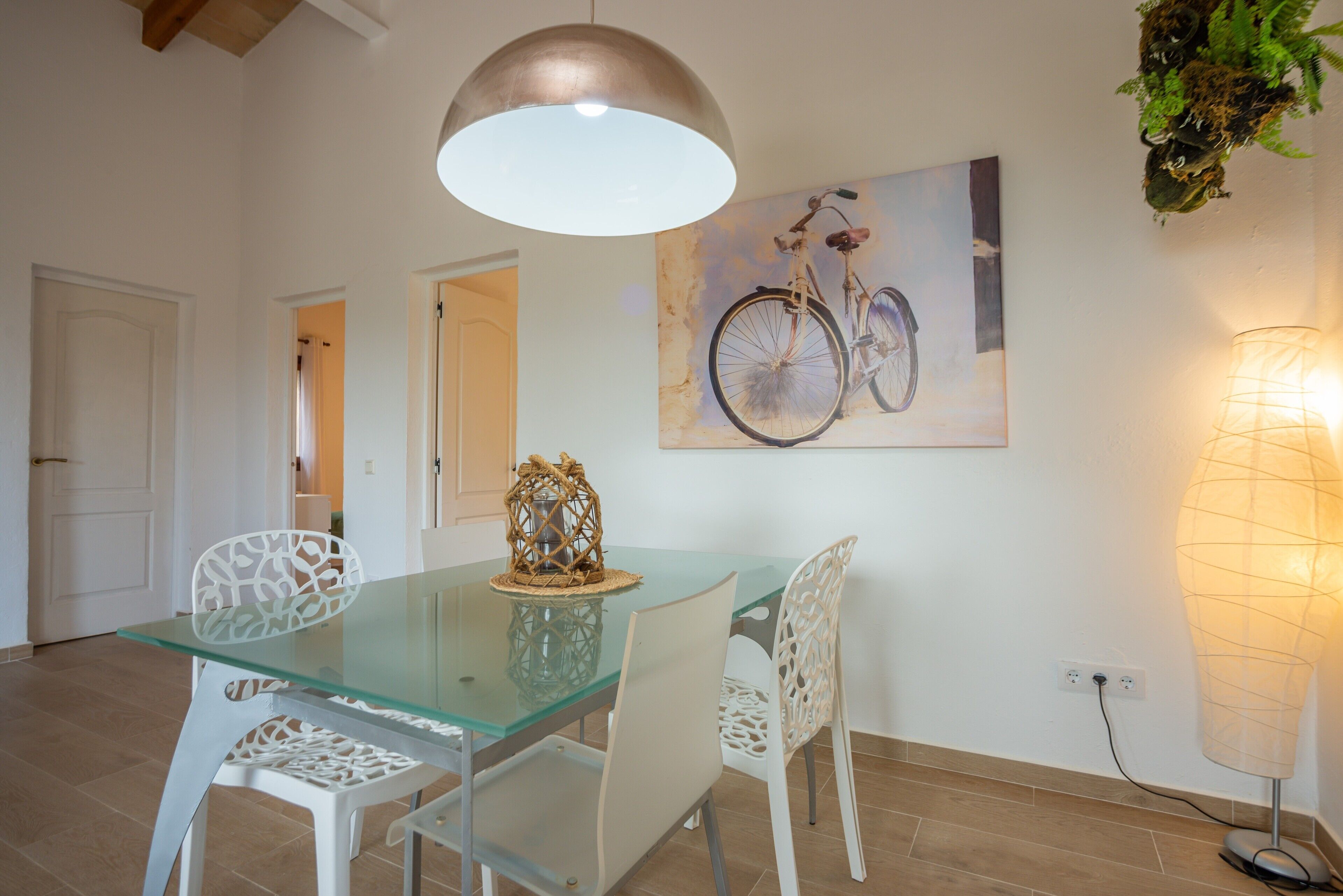 Un comedor con una mesa de vidrio, sillas blancas, una lámpara colgante y un cuadro de una bicicleta en la pared.