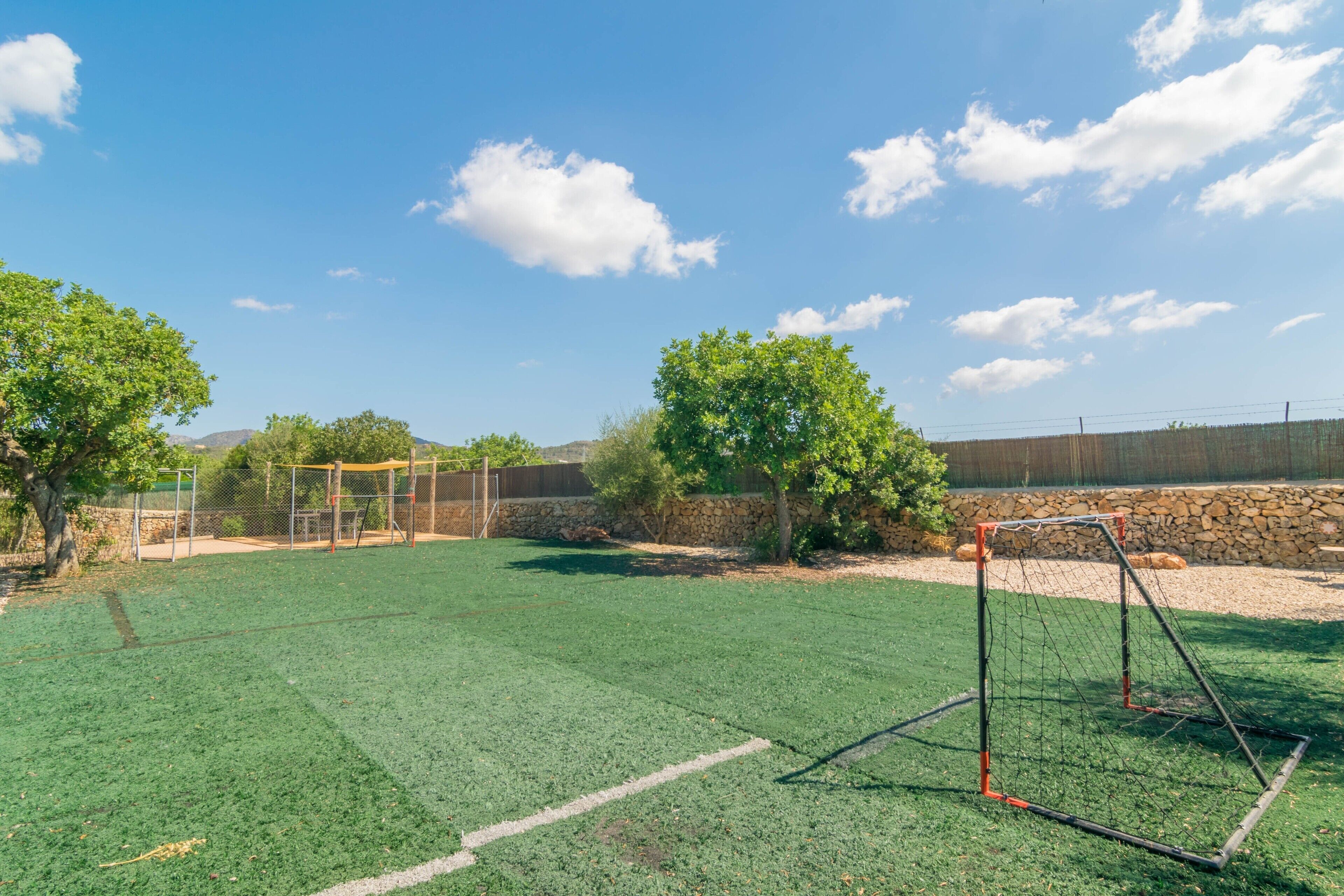 Un terrain de football avec des buts, entouré d’arbres et d’un mur de pierre.