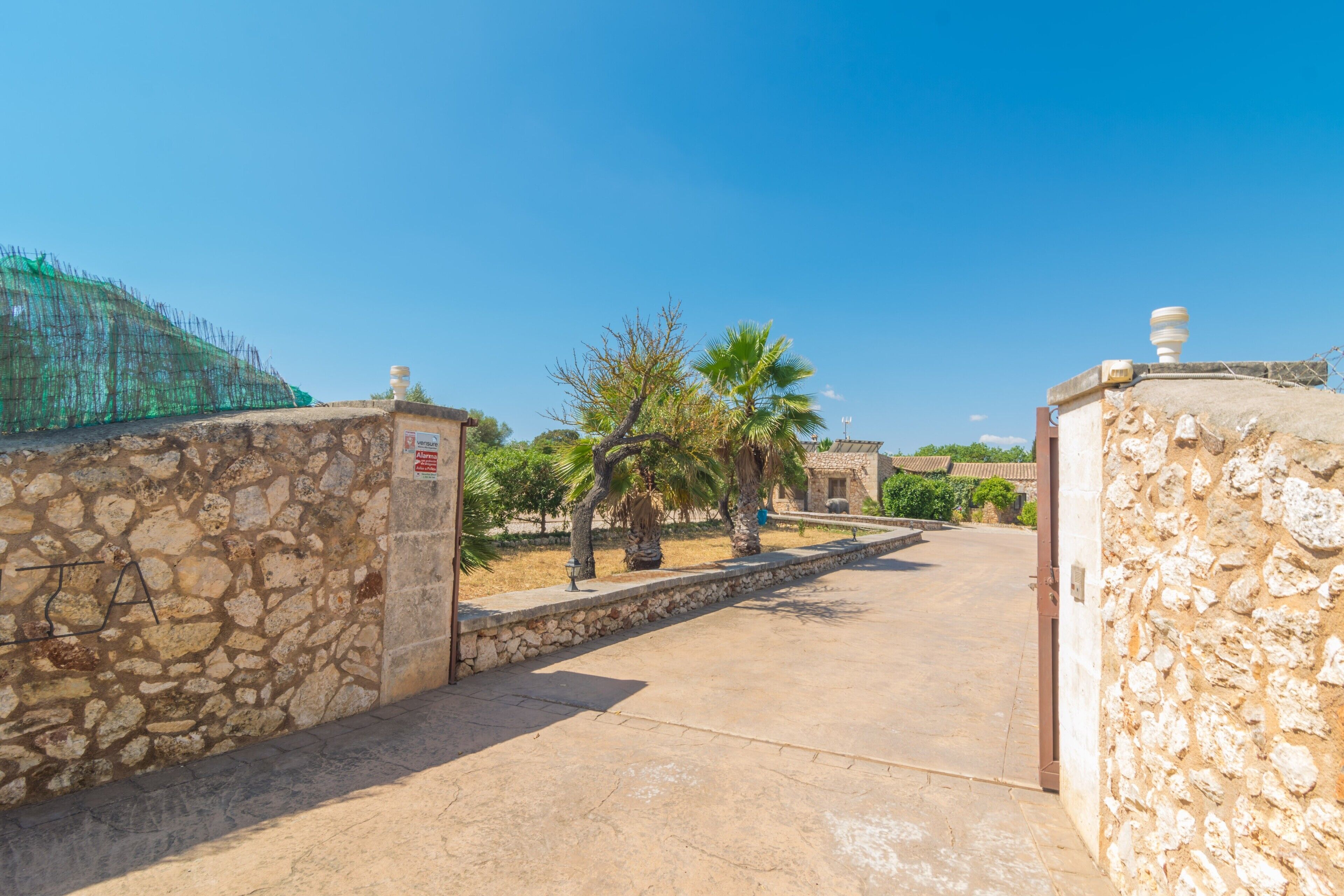 A stone-walled entrance with a clear sky, palm trees, and a traditional building in the background.