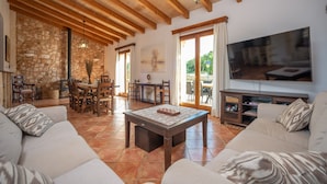 A spacious living room with a stone wall, wooden beams, a large TV, and a dining area with chairs and a table. - SA PUNTA - Villa with private pool in Son Carrio. Free WiFi (Sant Llorenç des Cardassar)