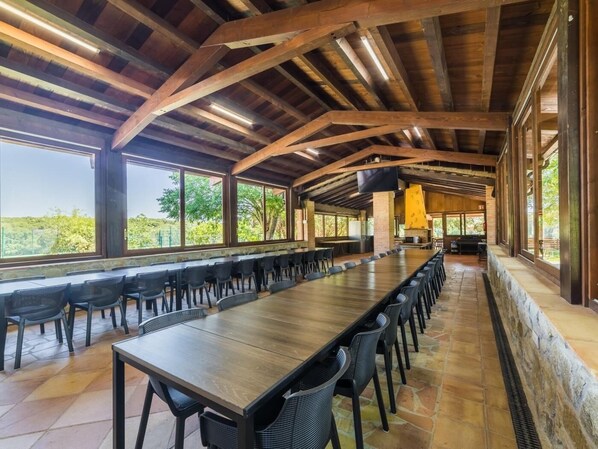 A long dining table with chairs, wooden beams, large windows, and a stone floor.