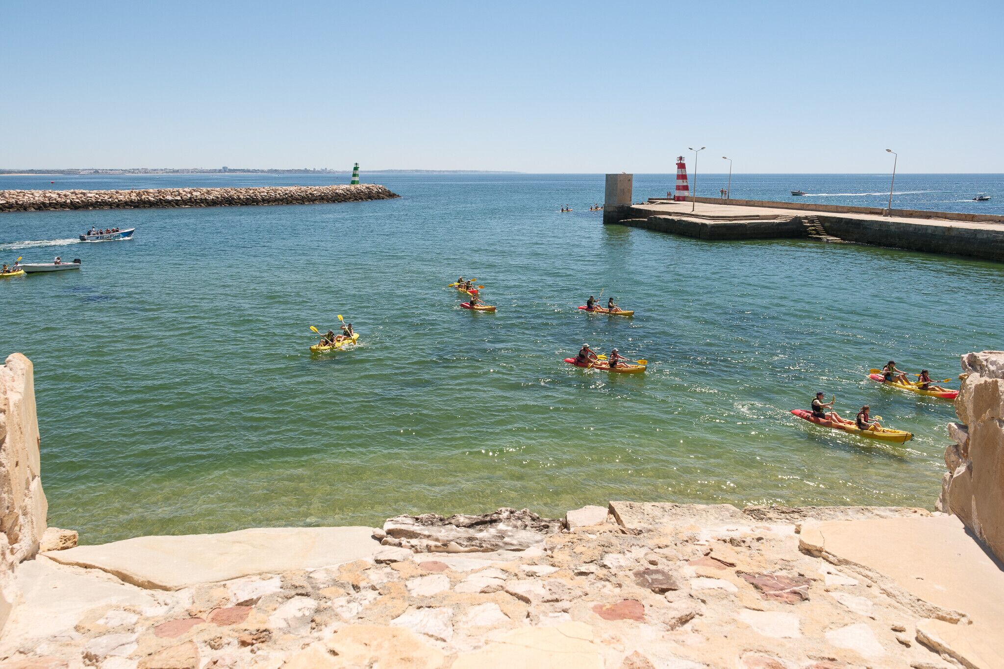 A coastal scene with kayakers on the water, a lighthouse, and a stone pier.