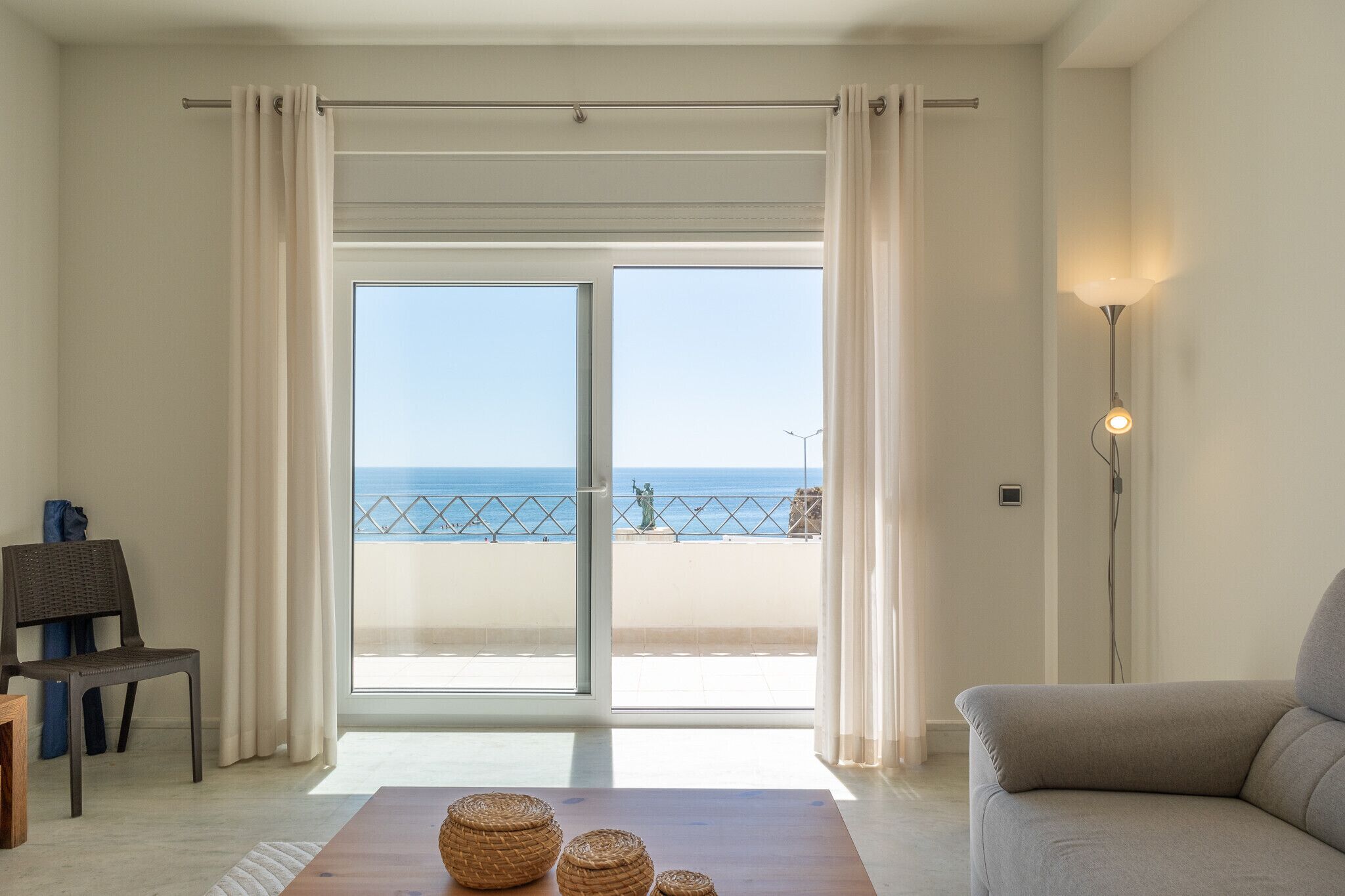 Een kamer met een grote schuifpui die uitkomt op een balkon met uitzicht op zee. Binnen in de kamer staat een bank en een stoel.