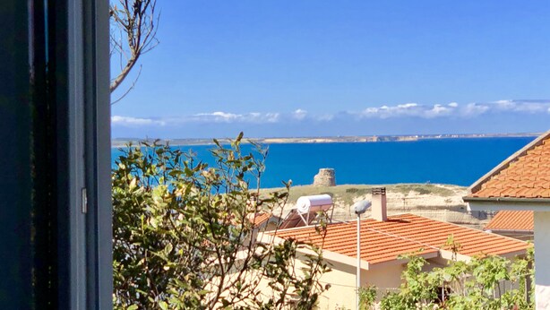 A coastal view through a window, featuring a clear blue sea, distant land, and a building with a tiled roof.