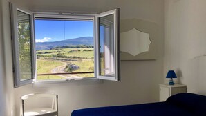 A bedroom with a bed, a chair, a nightstand with a lamp, and a window with an open view of a landscape.