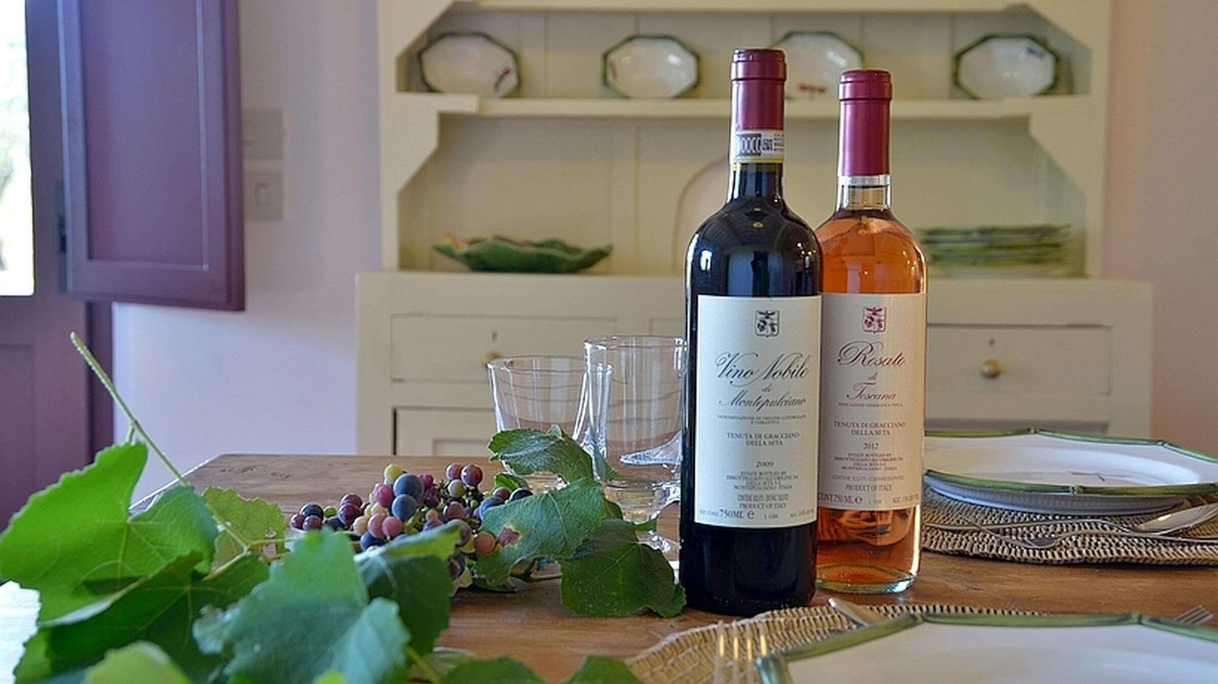 Two wine bottles on a wooden table with a glass and a bunch of grapes.
