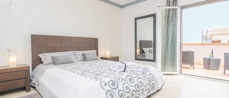 Une chambre dotée d’un grand lit, d’une table de chevet, d’un miroir et d’une porte-fenêtre coulissante donnant sur un espace extérieur aménagé avec du mobilier.