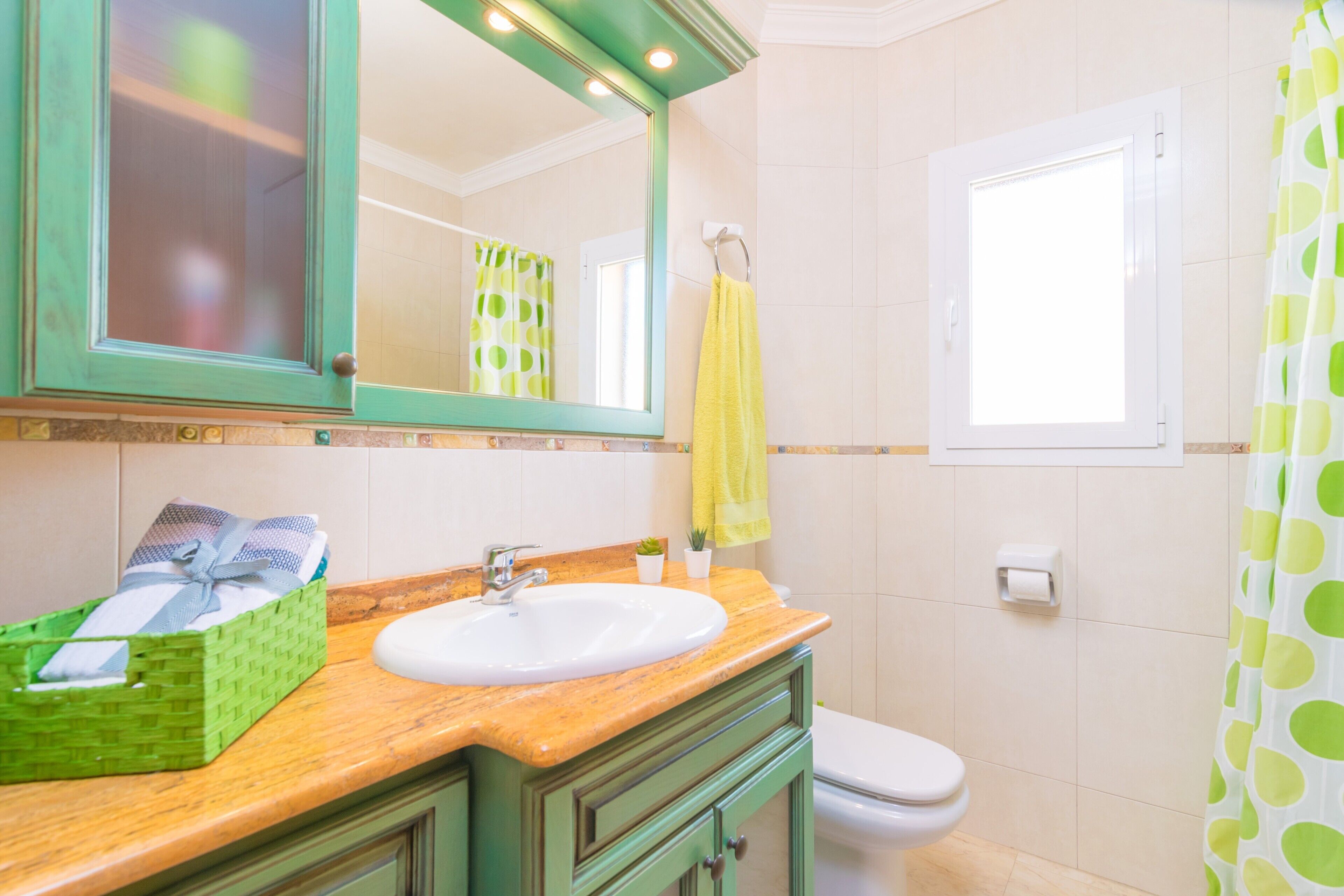 Une salle de bain avec un meuble-lavabo en bois, un lavabo blanc, un meuble vert, un miroir, une serviette jaune, un rideau de douche vert à pois et des toilettes blanches.