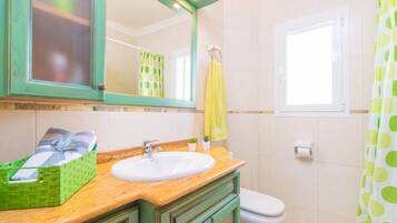 Une salle de bain avec un meuble-lavabo en bois, un lavabo blanc, un meuble vert, un miroir, une serviette jaune, un rideau de douche vert à pois et des toilettes blanches.