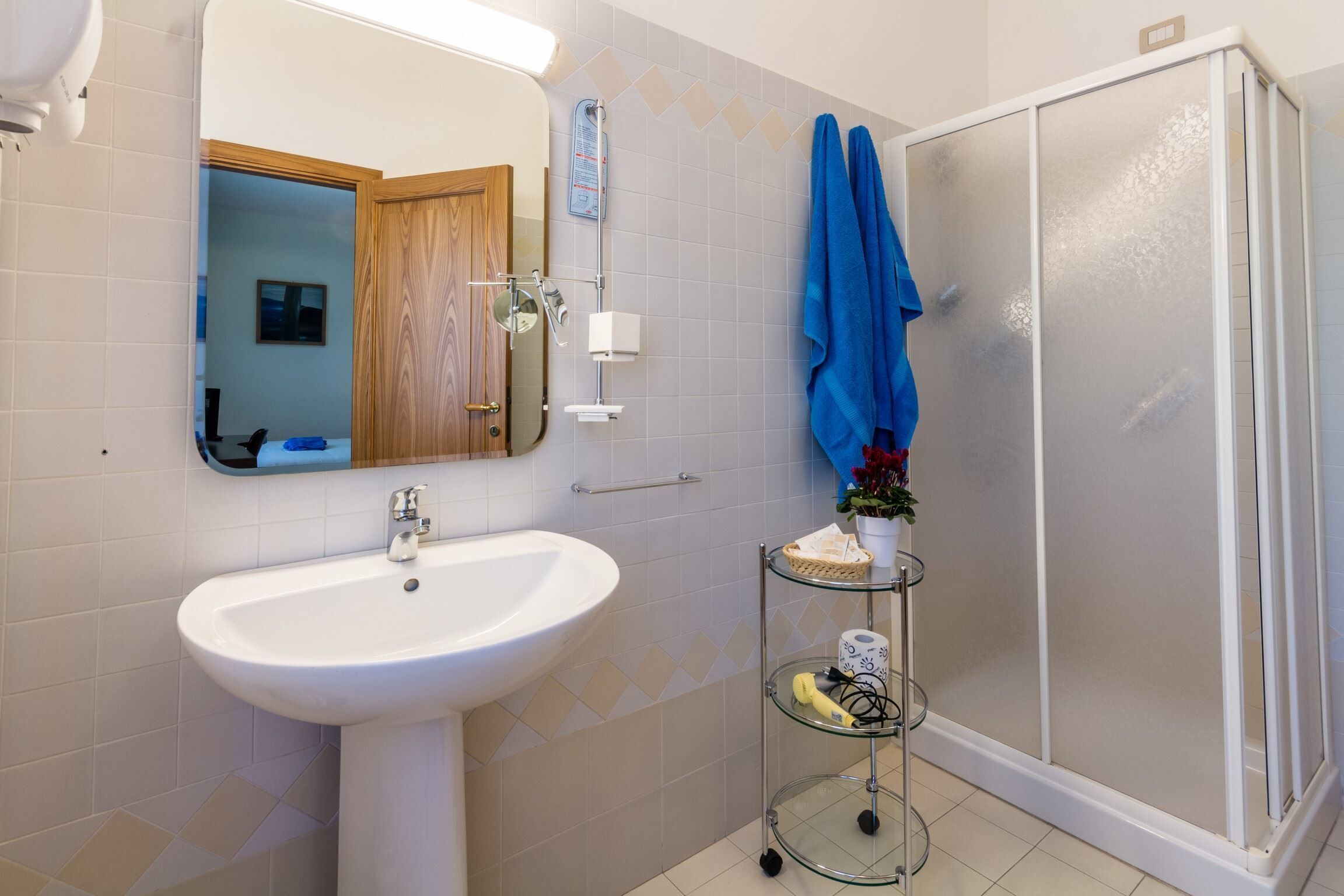 A bathroom with a white sink, a glass shelf with toiletries, a blue towel, and a glass shower enclosure.
