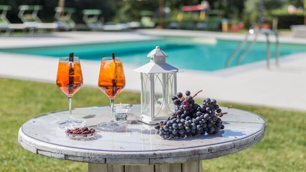 A table with two glasses of orange drink, a lantern, and a bunch of grapes by a pool.