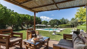 An outdoor patio with wooden furniture, a glass table, and a pool in the background.