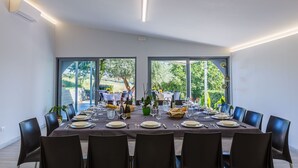 A dining area with a long table set for a meal, large windows, and a view of greenery outside.