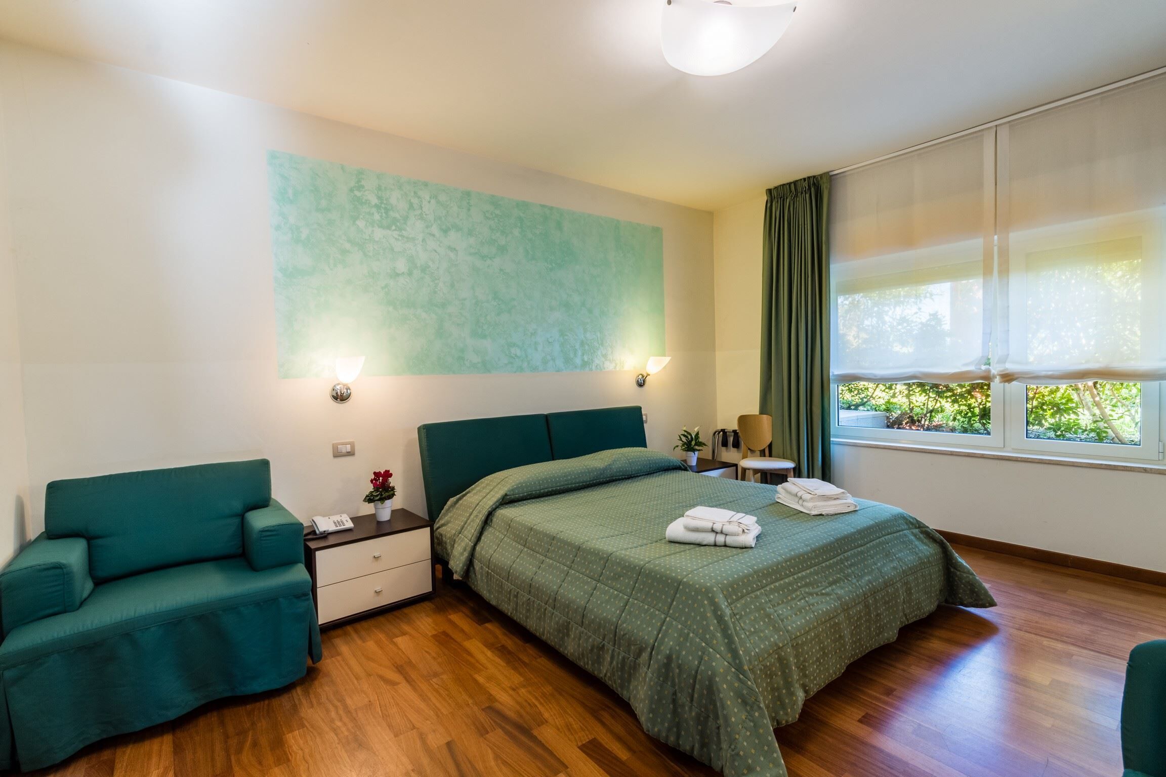 Un dormitorio con cama, sofá verde, mesita de noche, lámpara y ventana con cortinas.