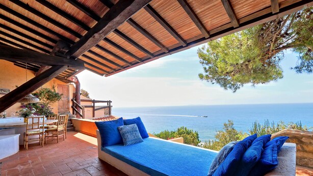 Eine Terrasse mit Holzbalkendecke, einer blauen Sitzgelegenheit mit Kissen, einem Tisch und Stühlen sowie Meerblick.