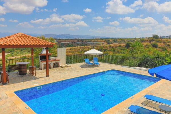 A swimming pool with lounge chairs and a pergola, surrounded by a stone deck and a view of distant hills.