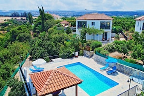 A swimming pool with a tiled deck, surrounded by greenery and a view of a hillside.