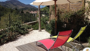 Een terras met twee lounge fauteuils, rood en geel, een witte parasol en een uitzicht op een berg en stad op de achtergrond.