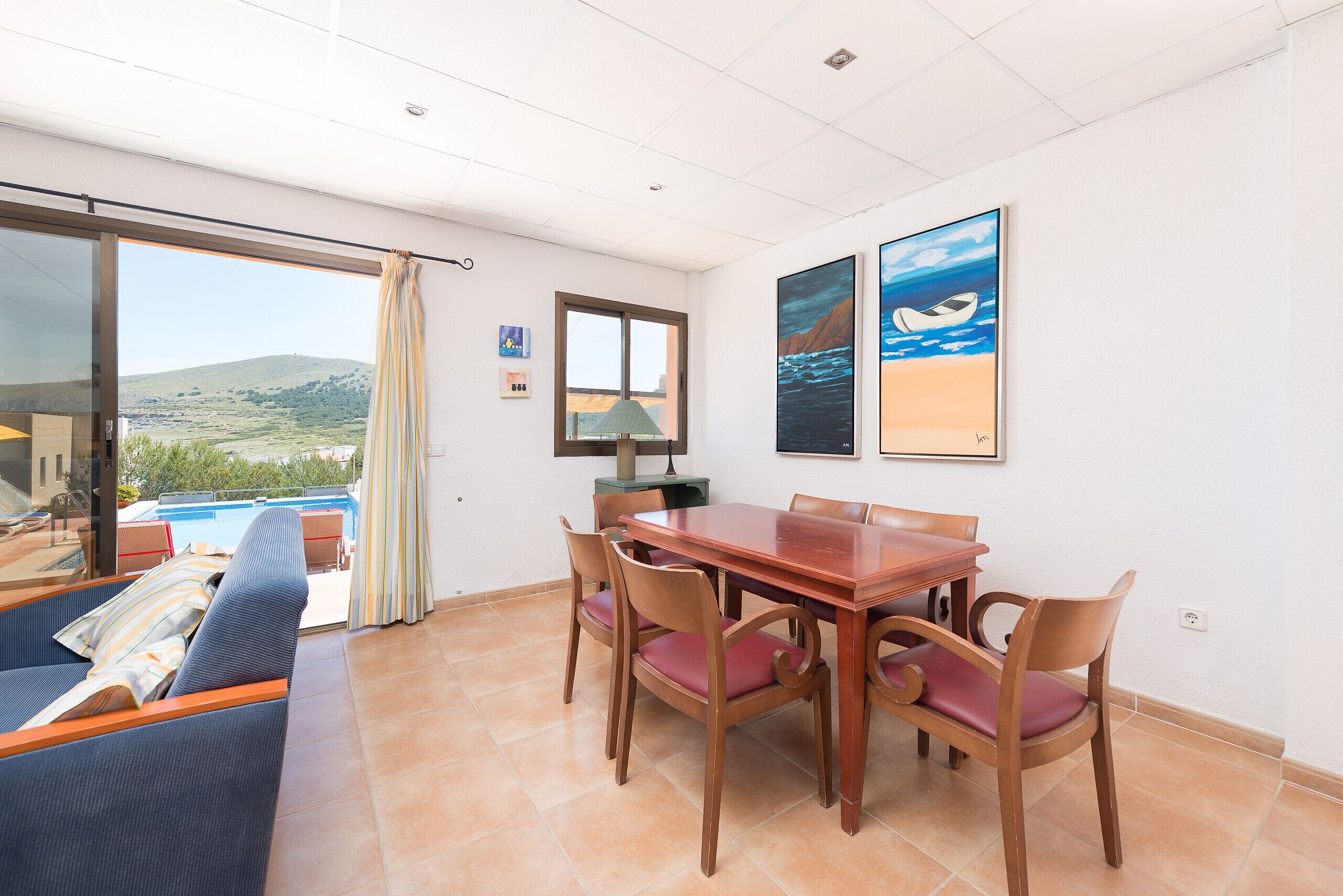 Un coin repas avec une table et des chaises en bois, un canapé et une vue sur une piscine et des collines.